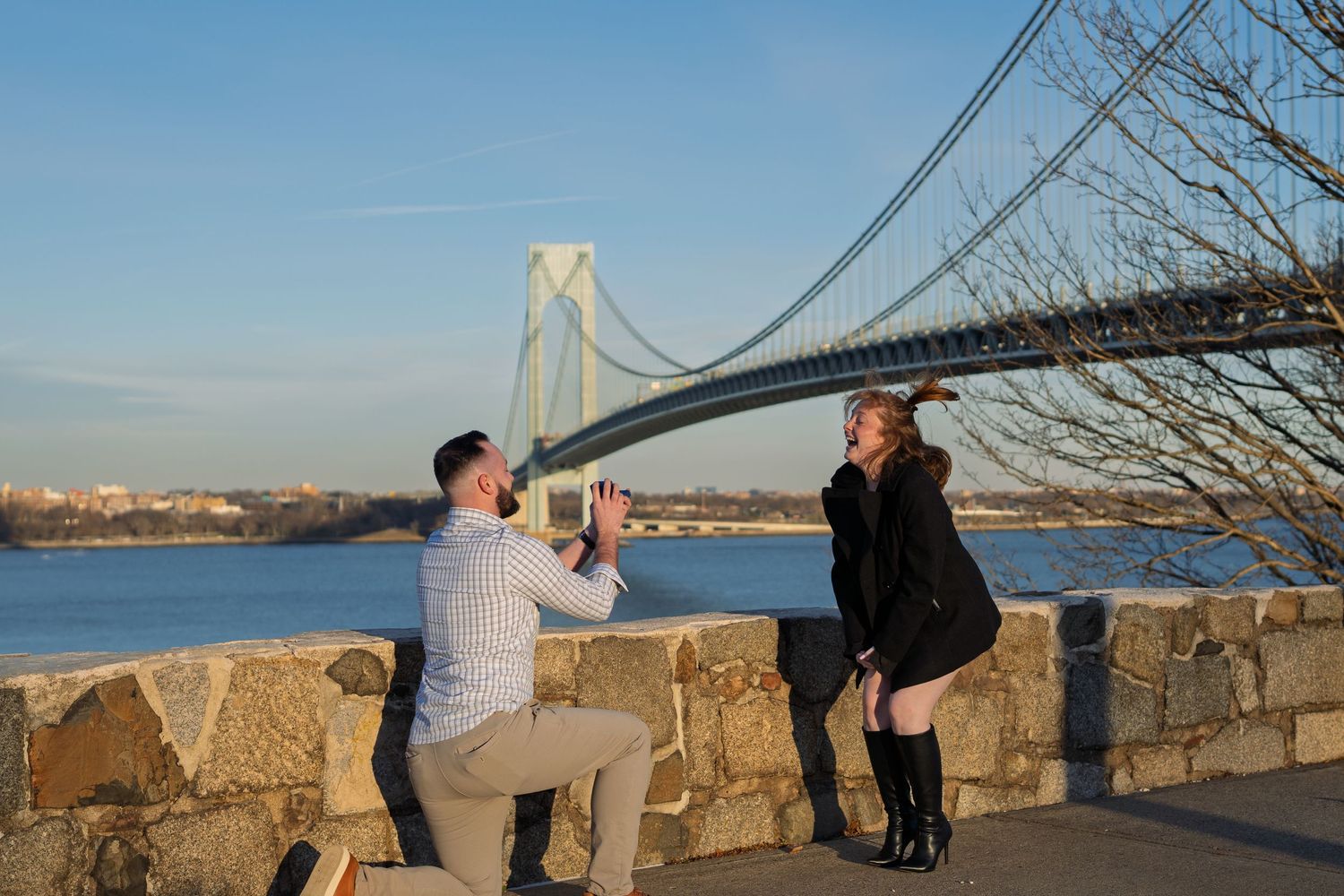 Romantic marriage proposal moment with a stunning suspension bridge and river in background.