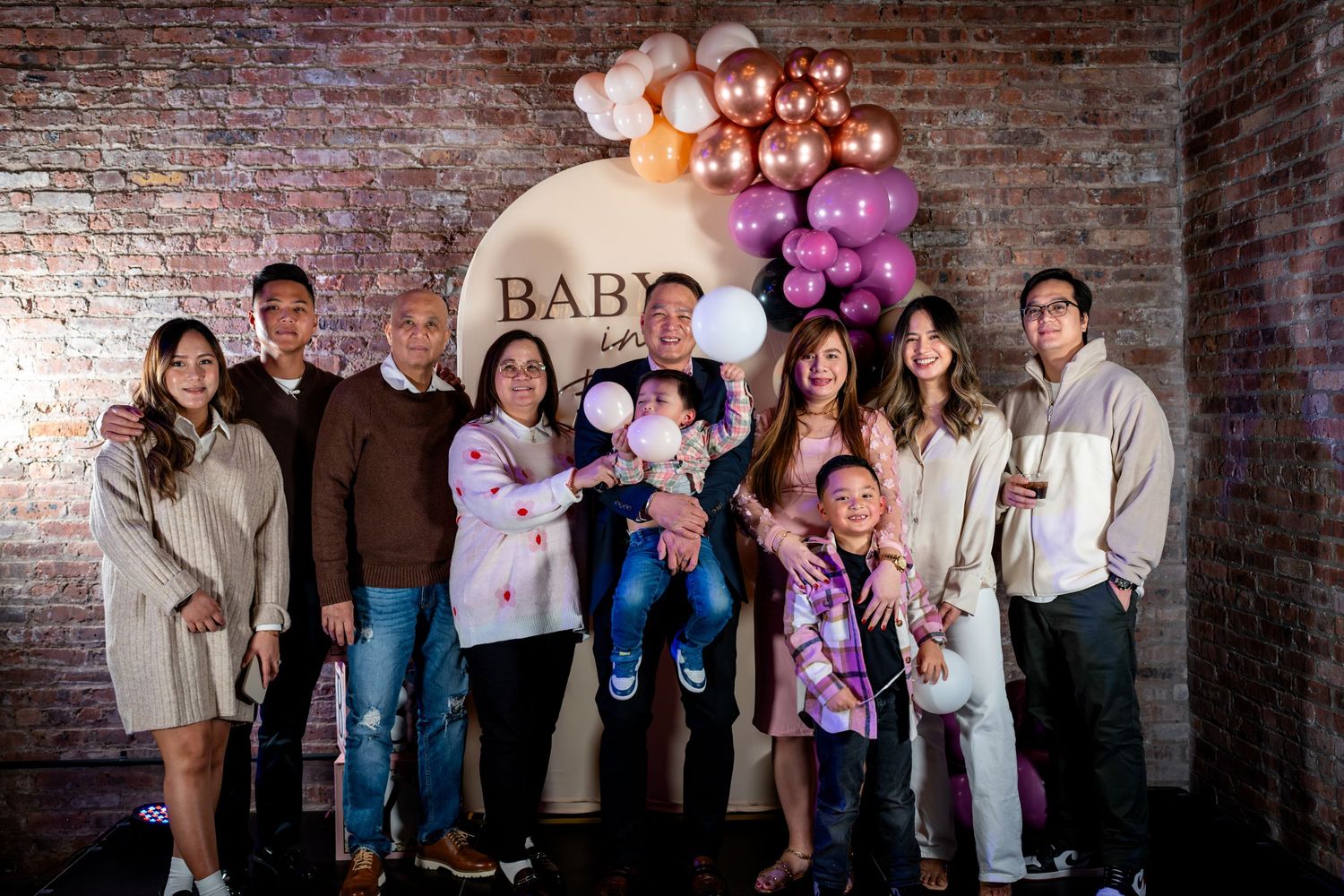 Group of eight family members pose together at baby shower celebration with balloon decor.