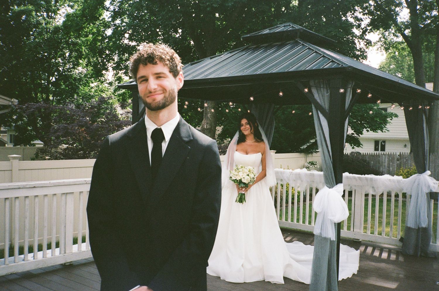 Groom in black suit smiles during outdoor wedding first look with bride in white gown near gazebo.