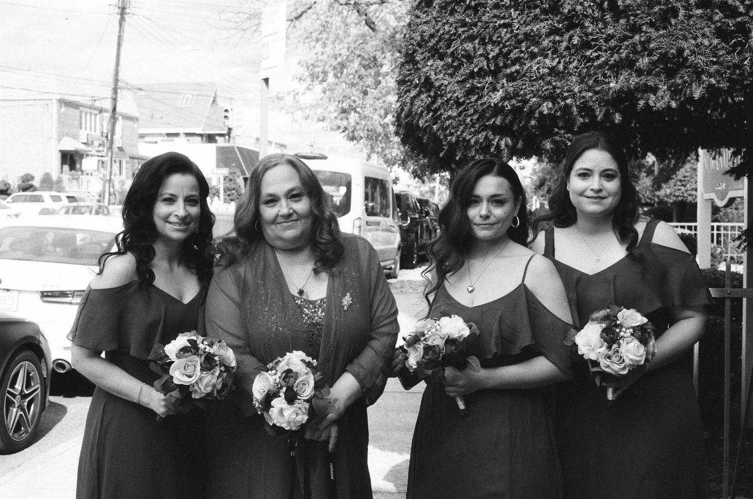Black and white photo of four bridesmaids in matching dresses holding white bouquets outdoors.