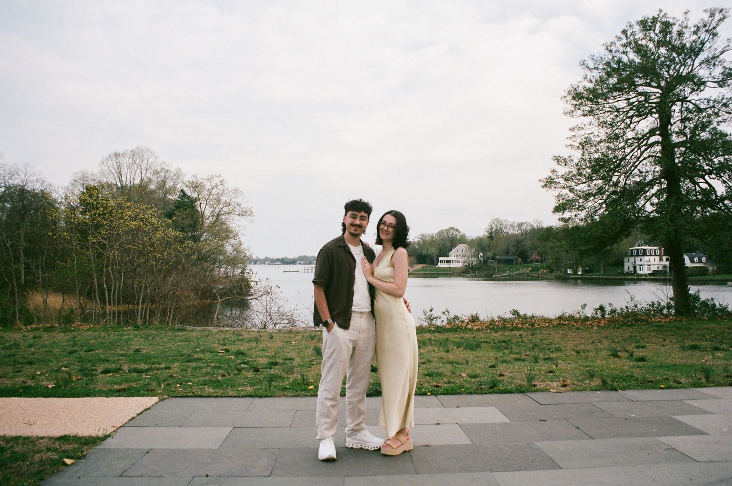 Smiling couple standing on a lakeside path, man in dark shirt and woman in light yellow jumpsuit.