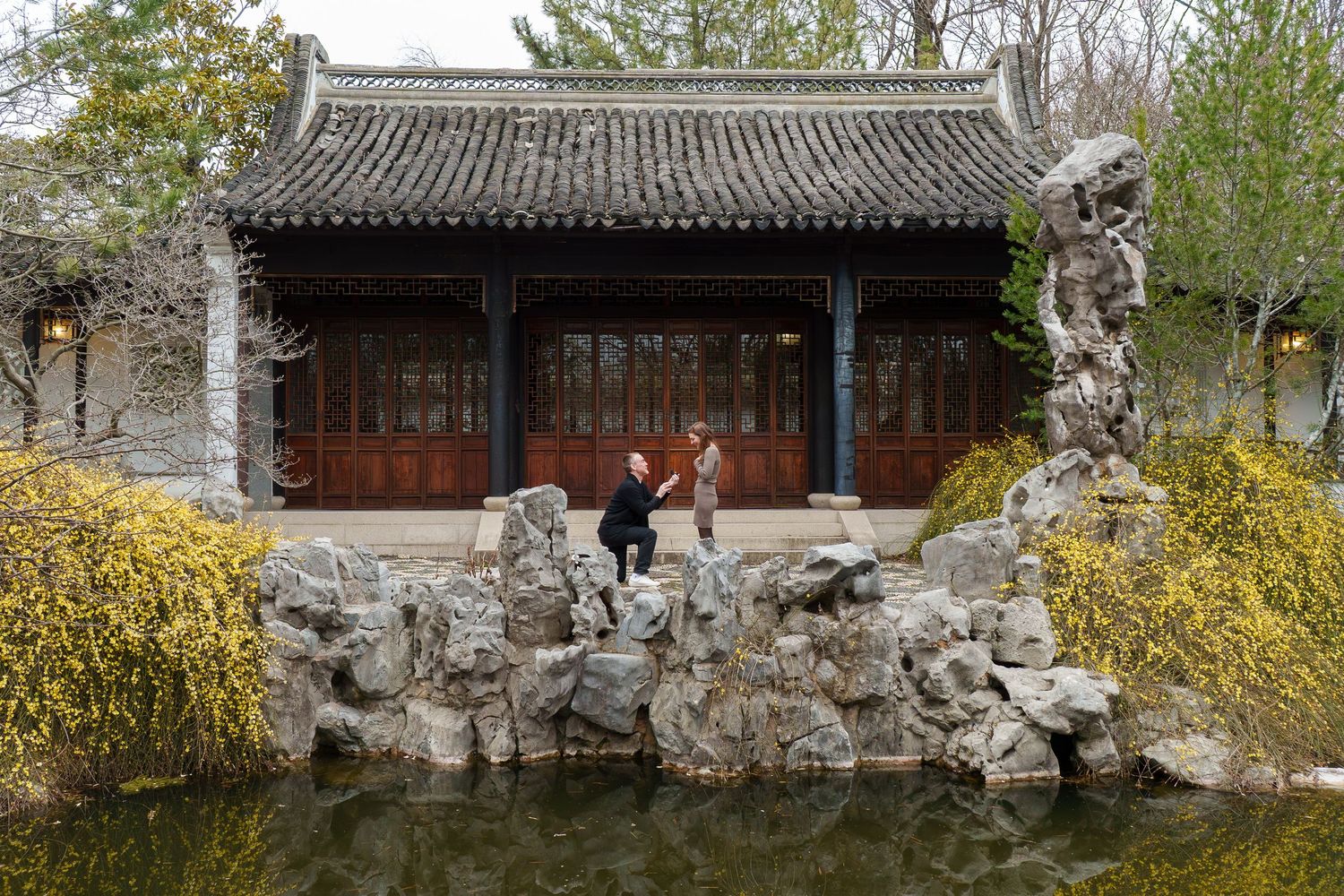 Couple standing on decorative rocks by a pond near a traditional Chinese pavilion.