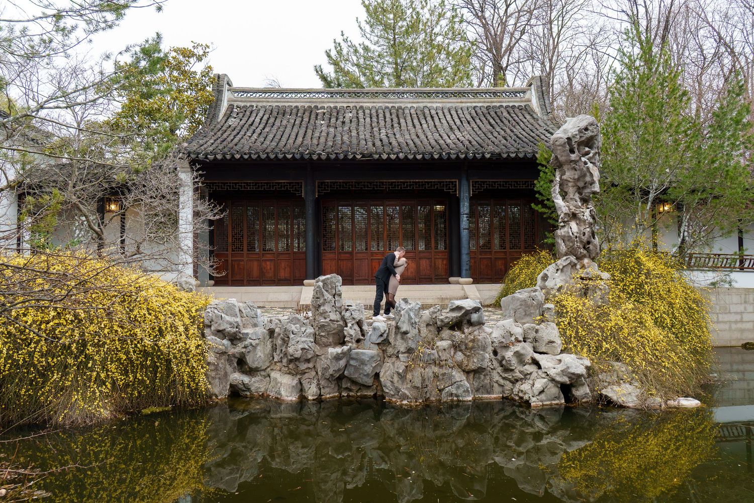Person posing on rocky landscape beside a reflective pond in front of a traditional Chinese garden pavilion.