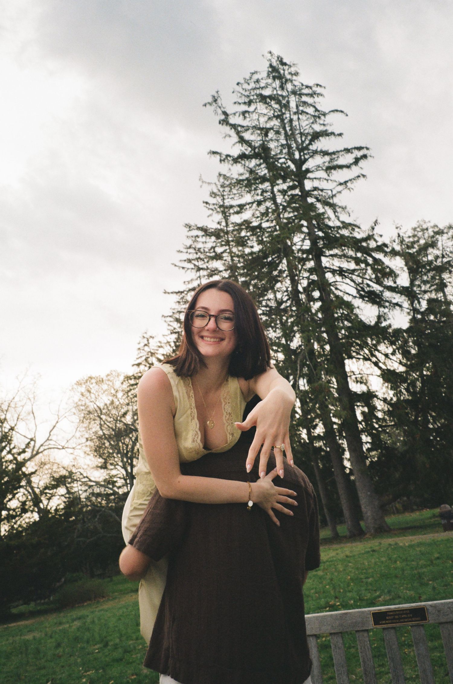 Smiling woman with glasses hugging her partner in a park with tall evergreen trees.