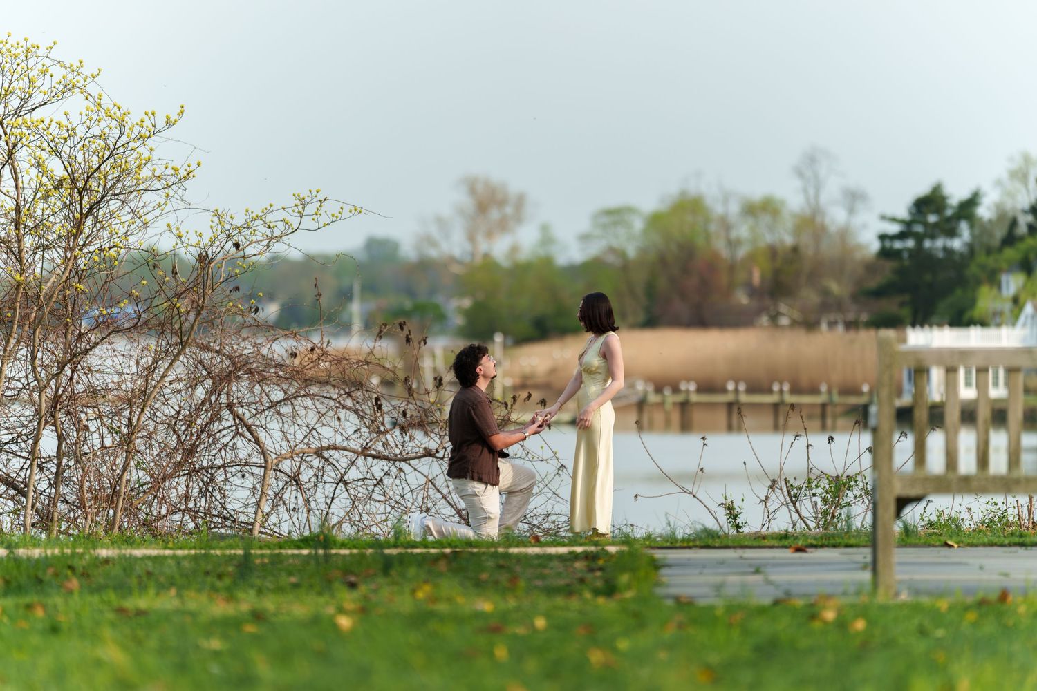 Couple holding hands during a romantic marriage proposal on a scenic waterfront walkway.