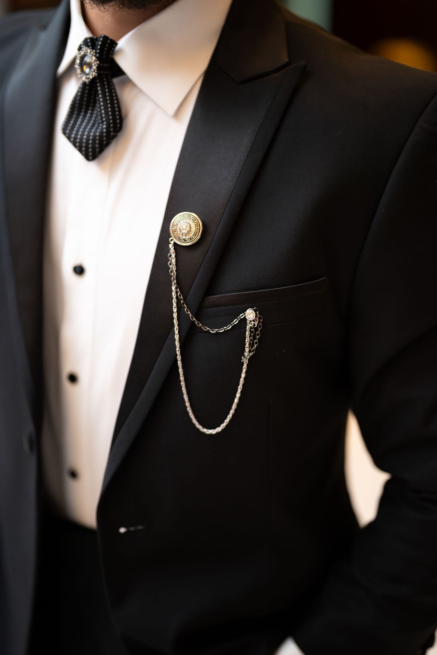 Groom wearing black tuxedo with gold lapel pin and chain accessory detail closeup.