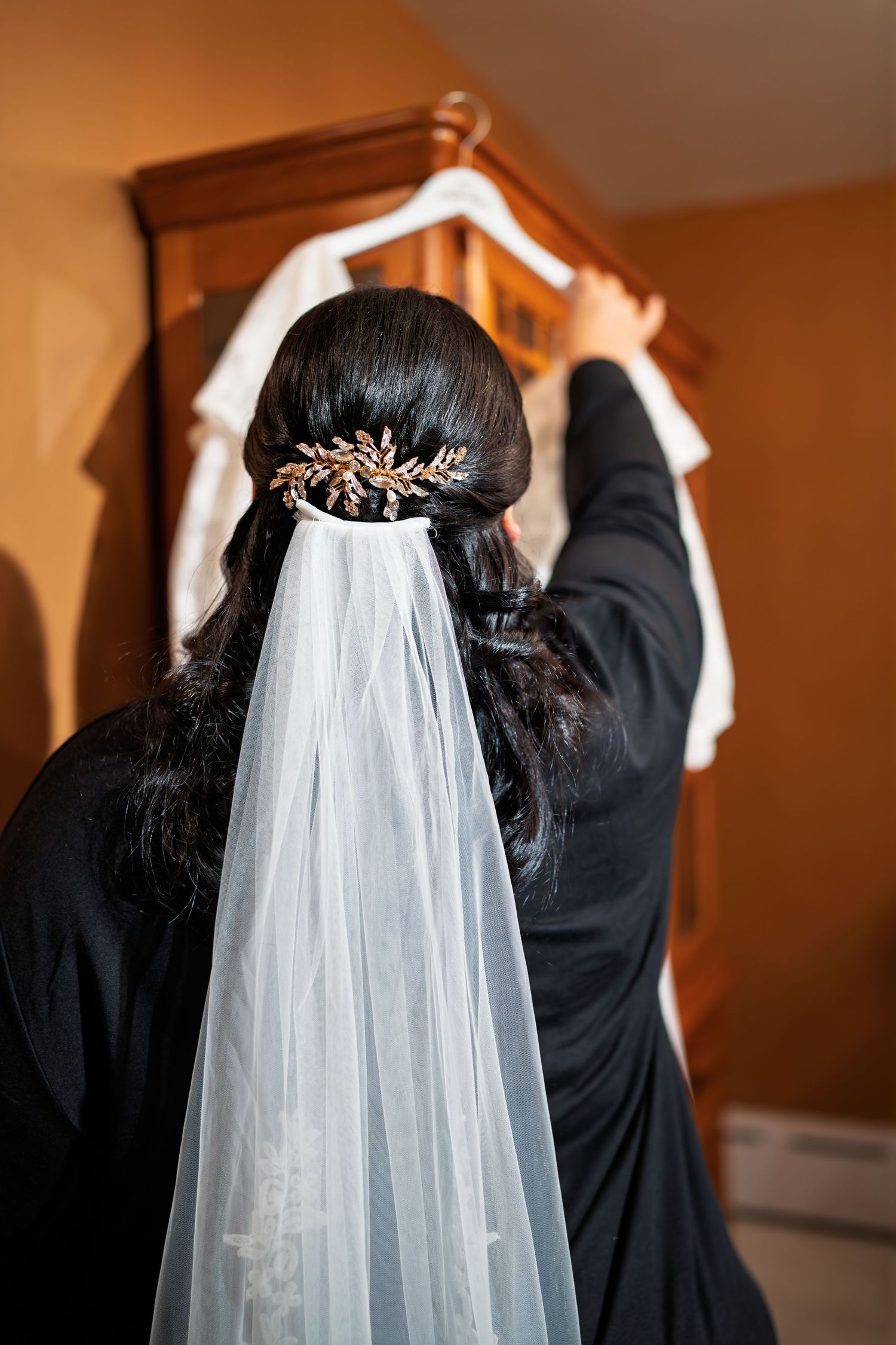 Bride with veil and gold hair accessory reaching for her wedding dress on a wooden armoire.