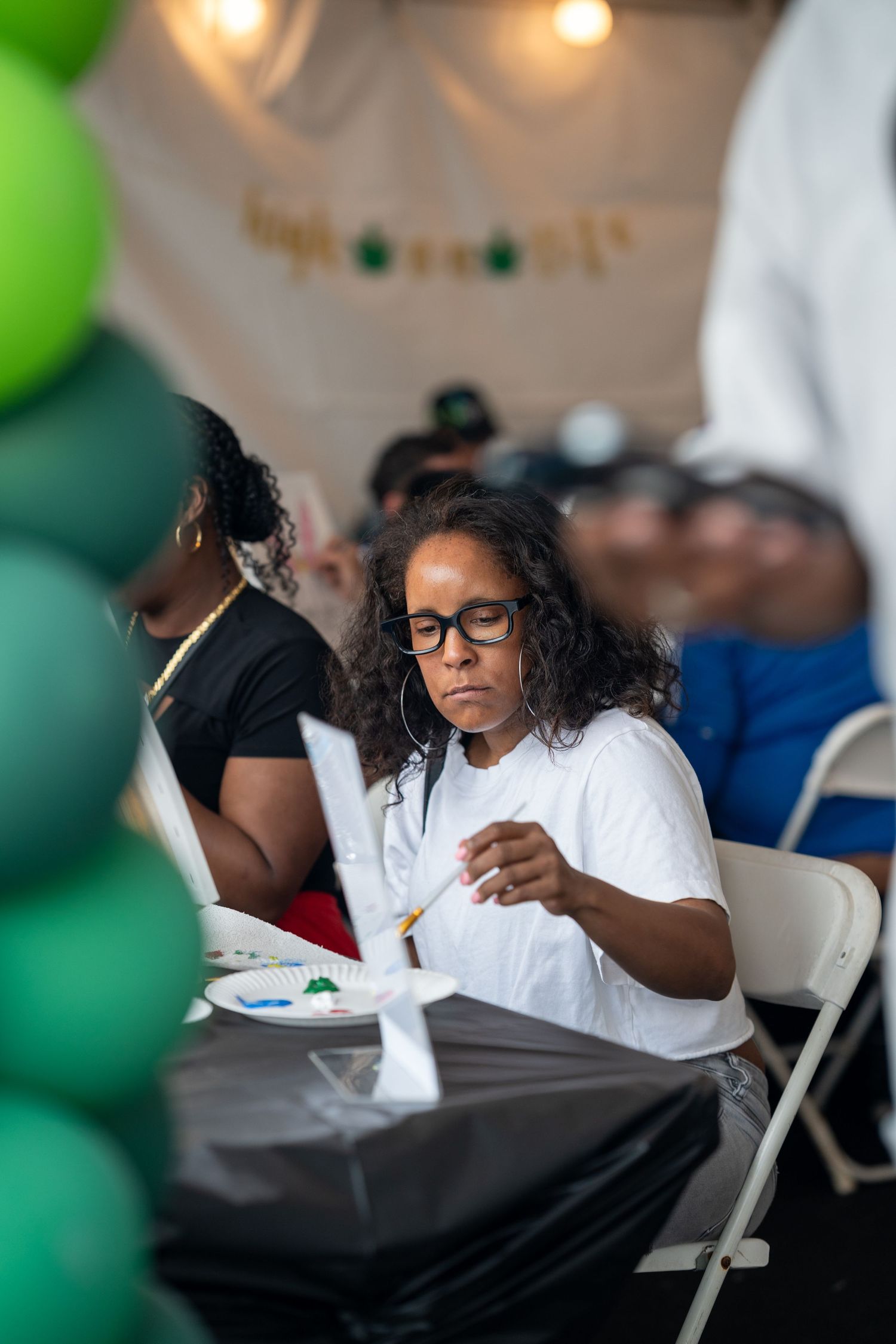 Woman with glasses painting at an outdoor art event with green balloons and festive decorations.