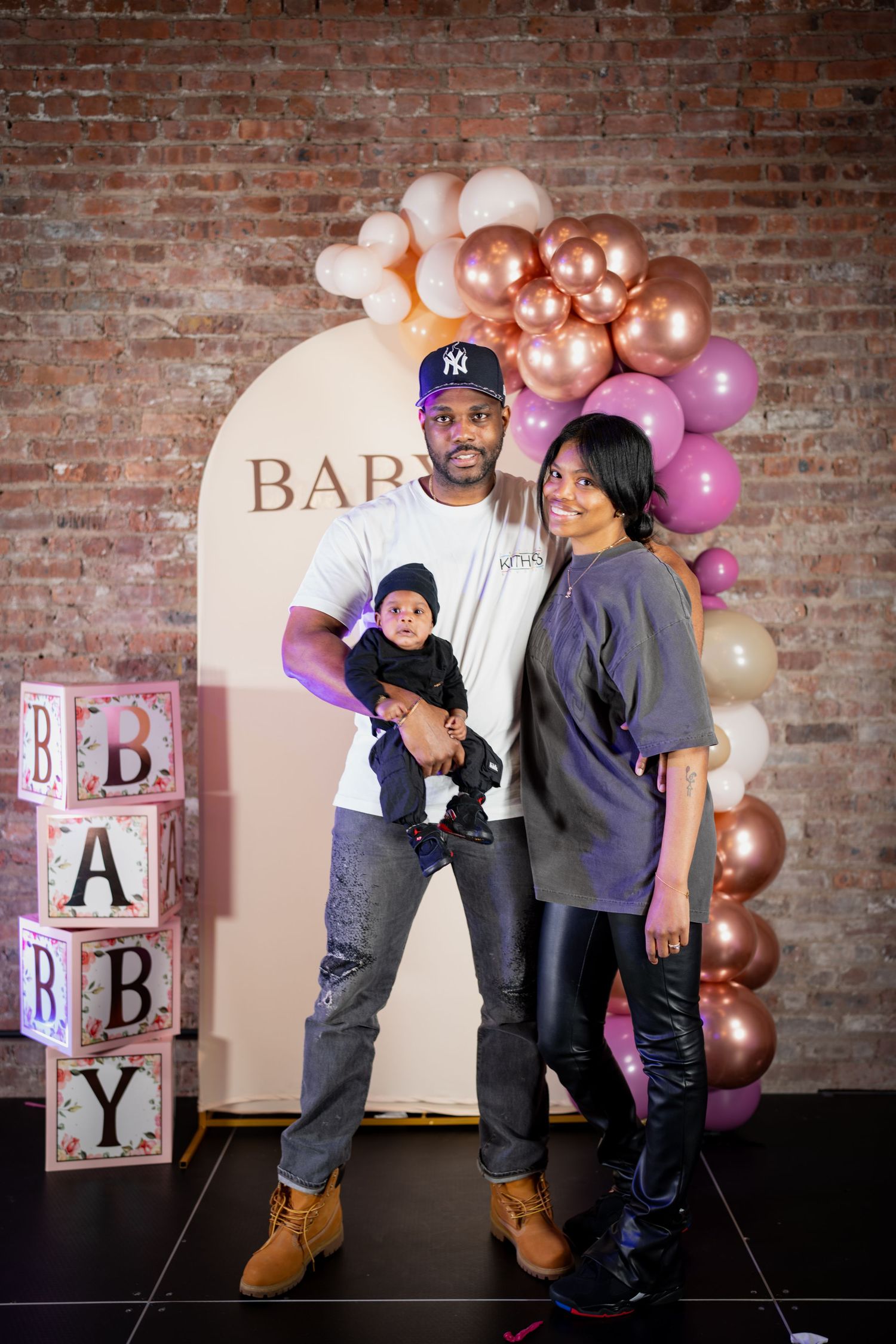 A man holding a baby and a woman posing together in front of a baby shower balloon arch backdrop.