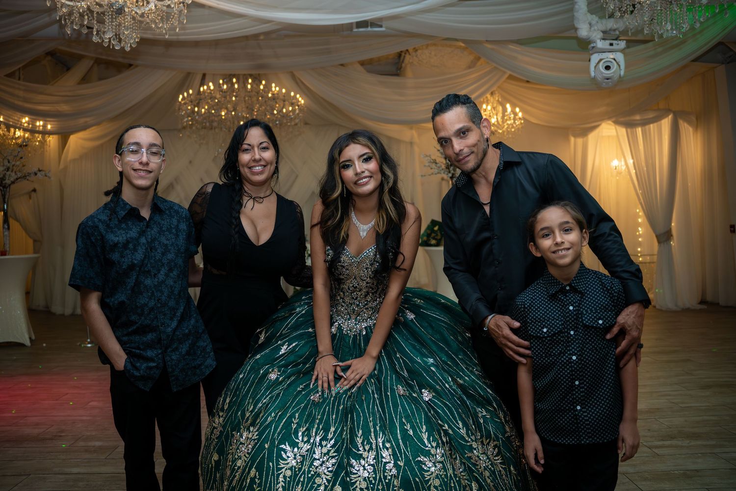 Family of five poses together in formal attire at a quinceañera celebration in decorated venue.