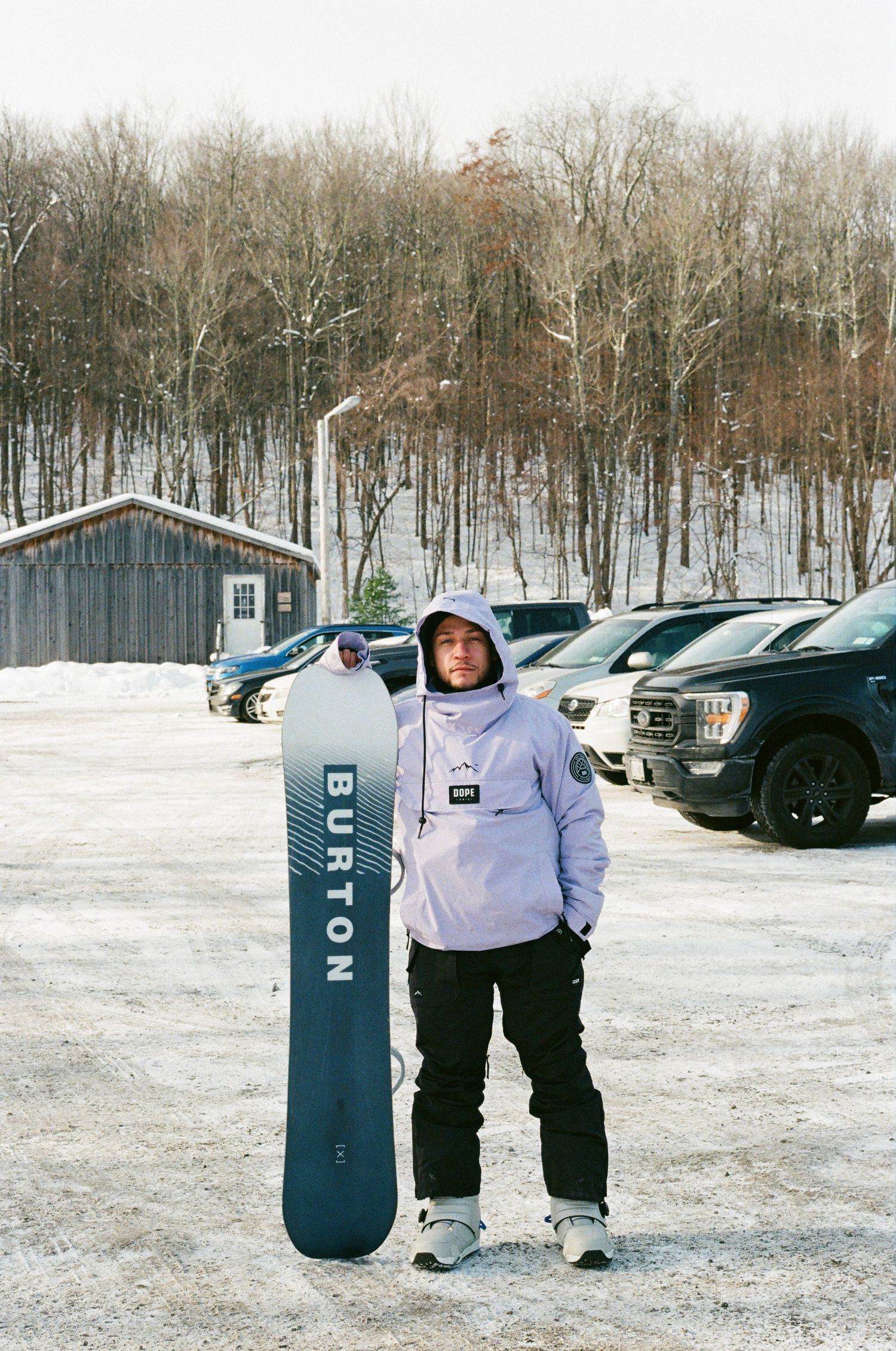 Person in purple hoodie holding Burton snowboard in snowy parking lot surrounded by trees.