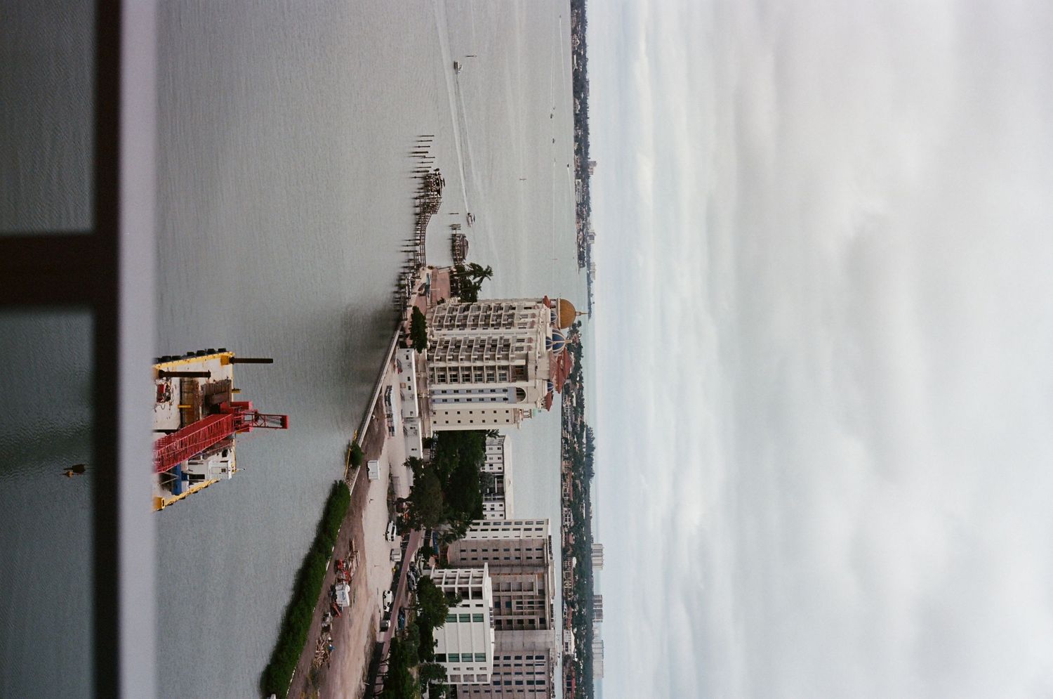 Aerial waterfront city view showing buildings along a harbor with cranes and overcast skies.