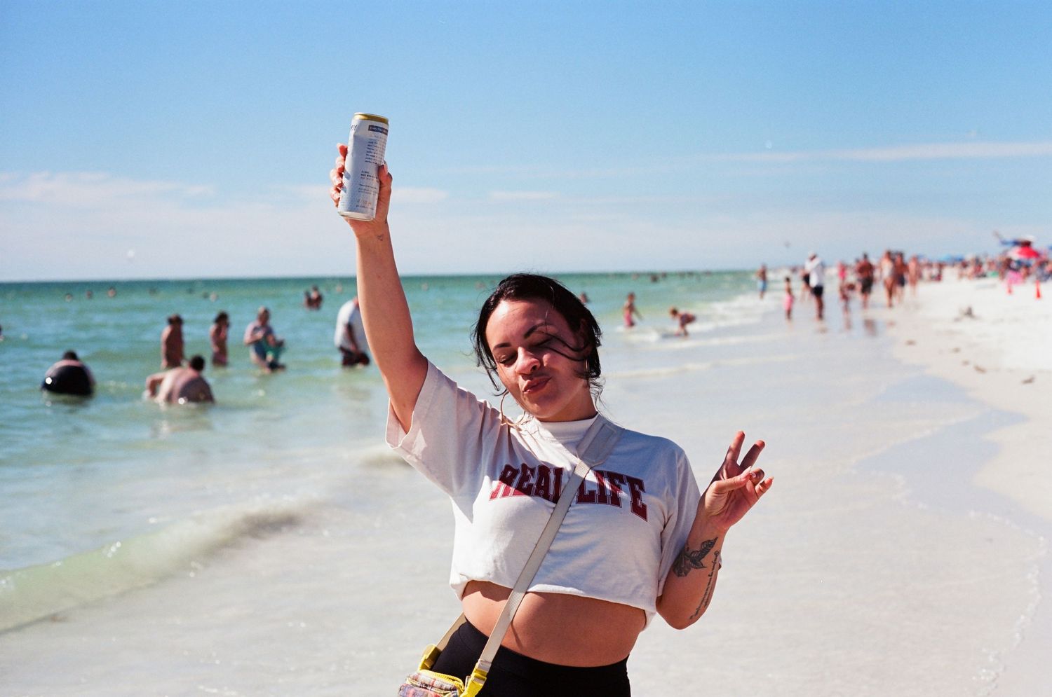 Young woman in white crop top holding a can up at a sunny crowded beach shoreline.