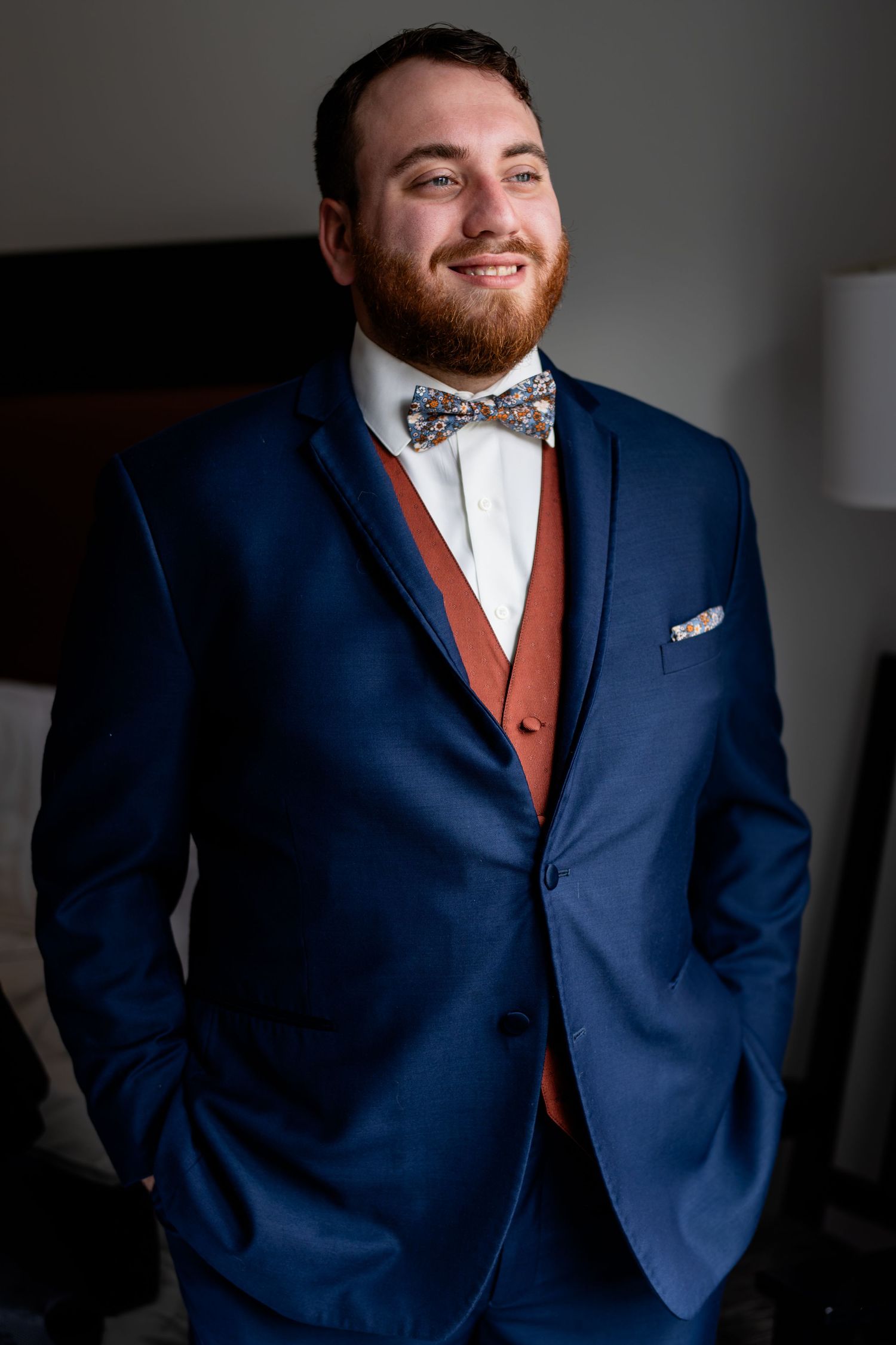 Handsome groom in navy blue suit with rust vest and floral bow tie smiles confidently indoors.
