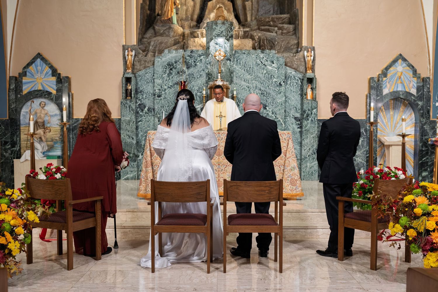 Wedding ceremony at ornate church altar with bride, groom, and priest surrounded by floral arrangements.
