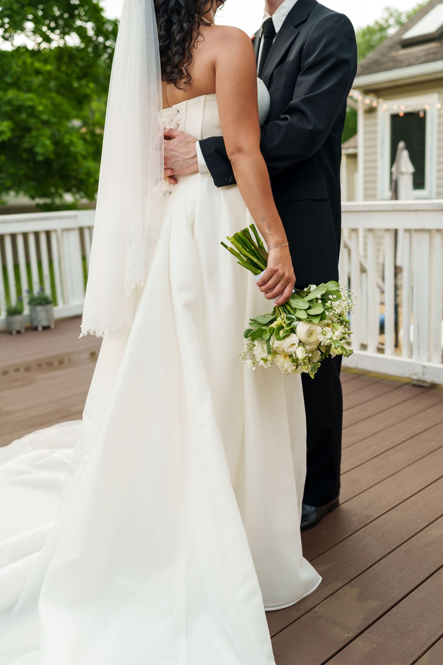 A bride in a white gown and veil holds a bouquet while embracing her groom in a black suit on a wooden deck.