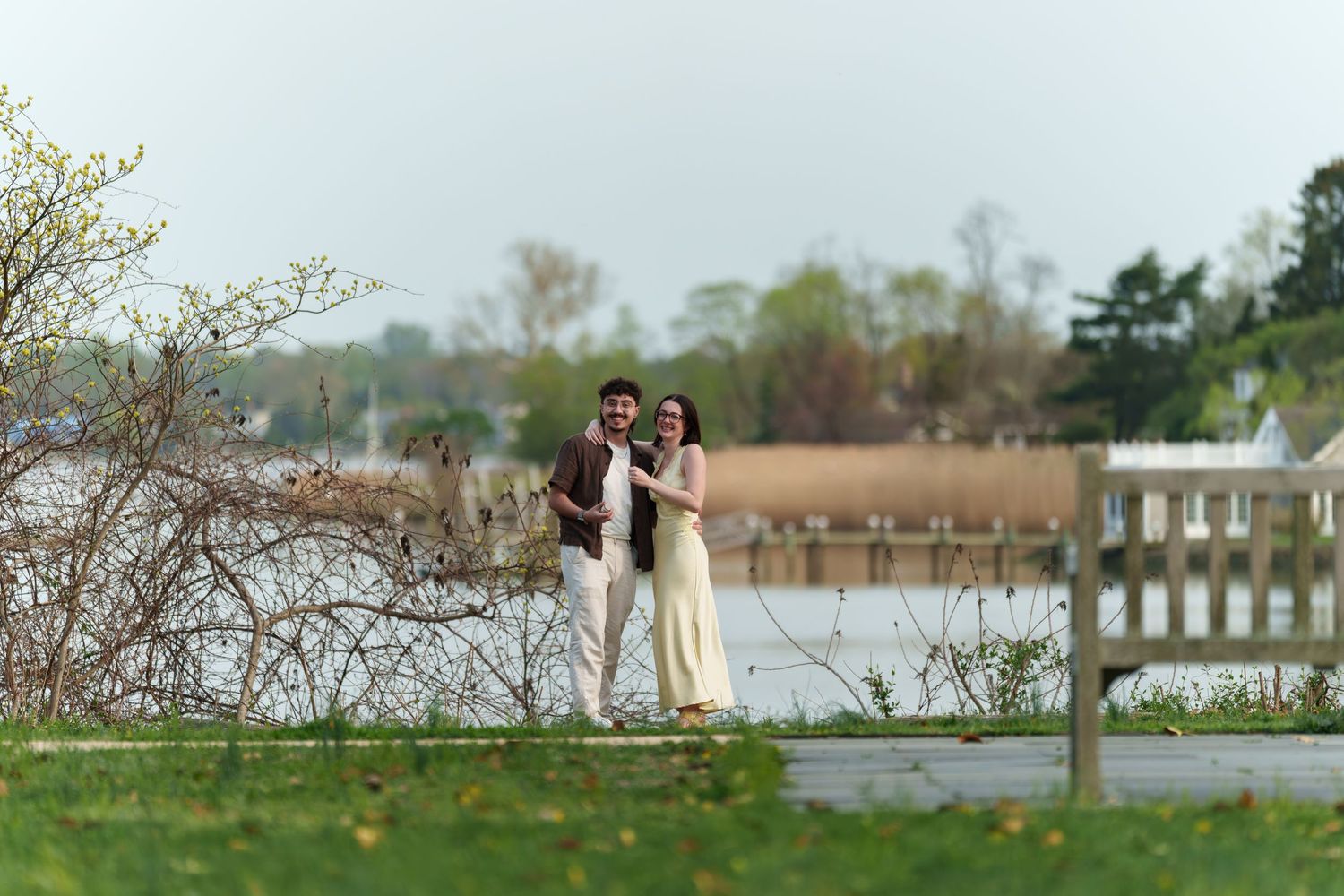 Happy engaged couple embracing and smiling near a tranquil waterfront with spring foliage.