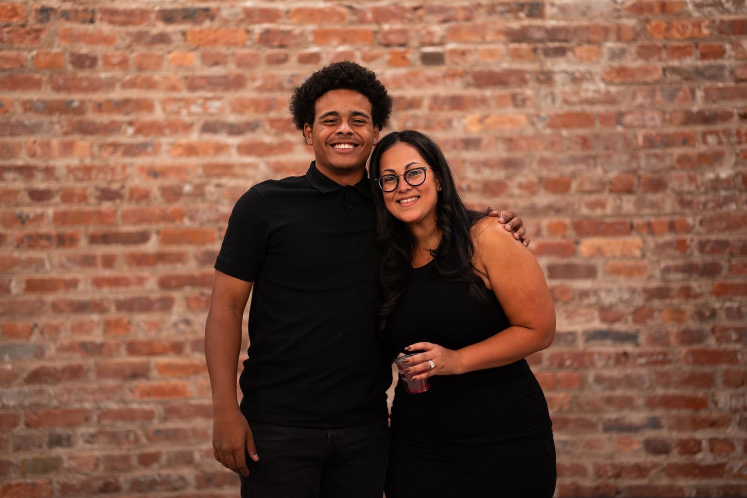 A young couple in matching black outfits smiling together against a rustic brick wall.