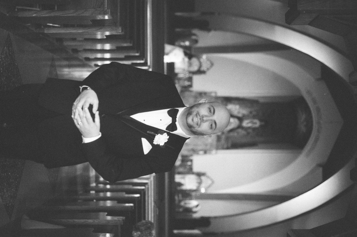 Elegant black and white portrait of a groom in a tuxedo standing inside a ornate church.