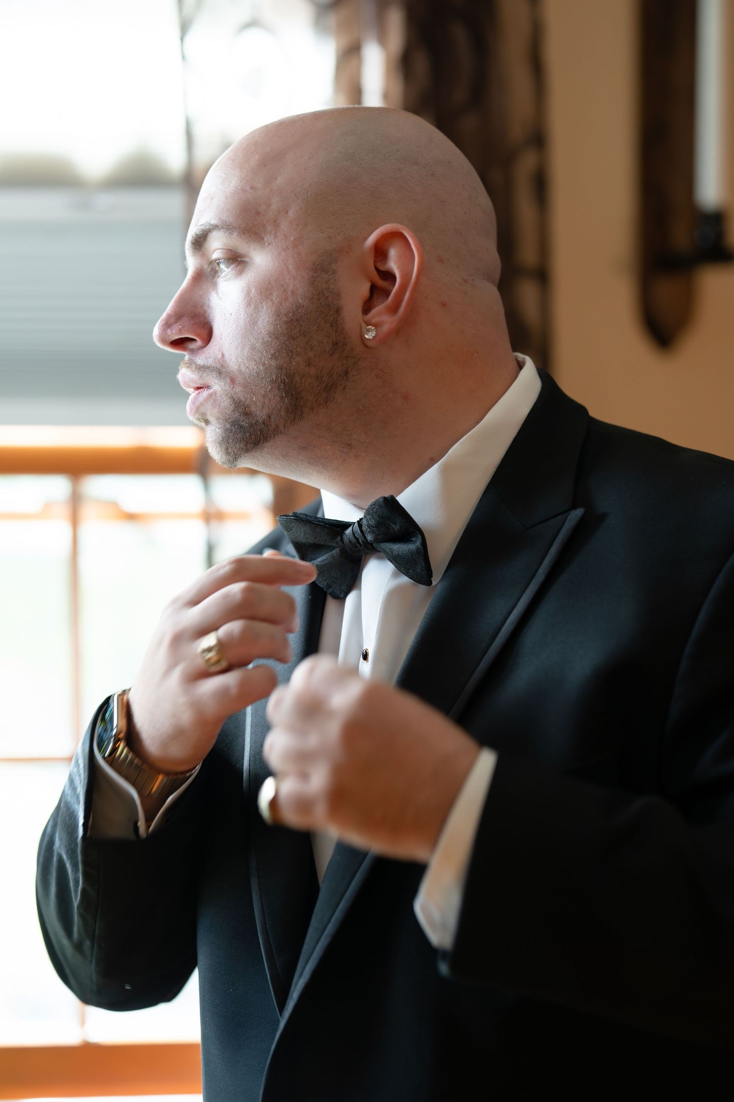 Dapper bald groom adjusting black bow tie in tuxedo before wedding ceremony.