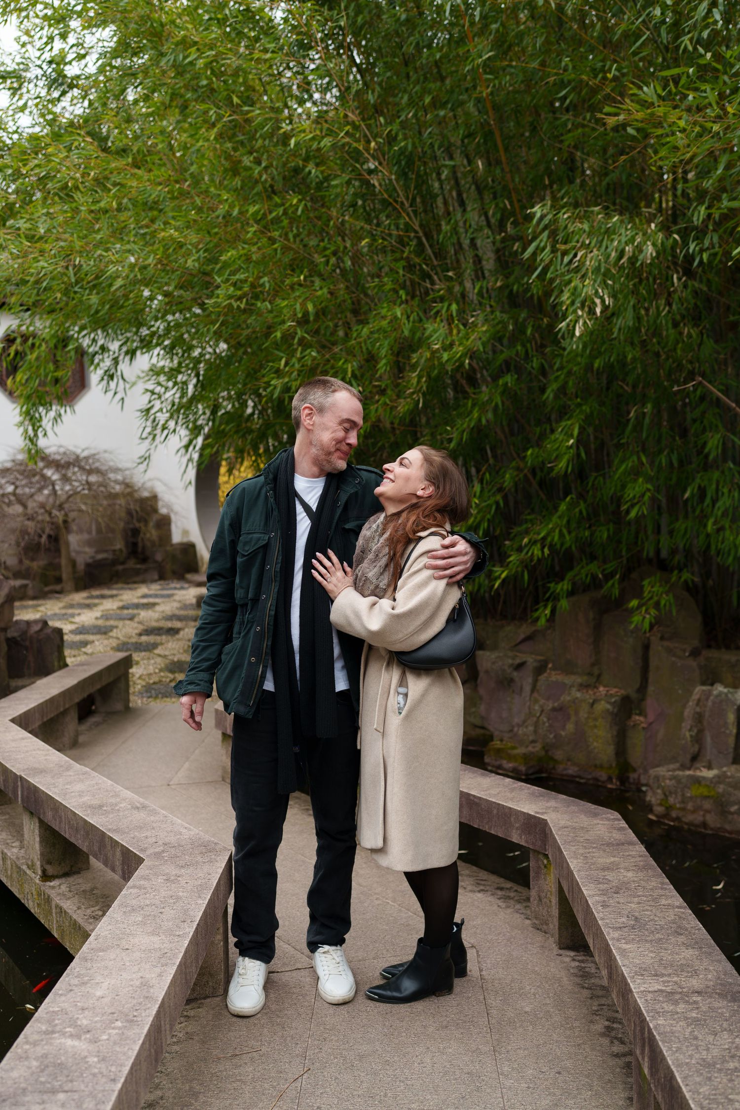 Smiling couple walking arm in arm on a stone bridge with tall green bamboo in the background.