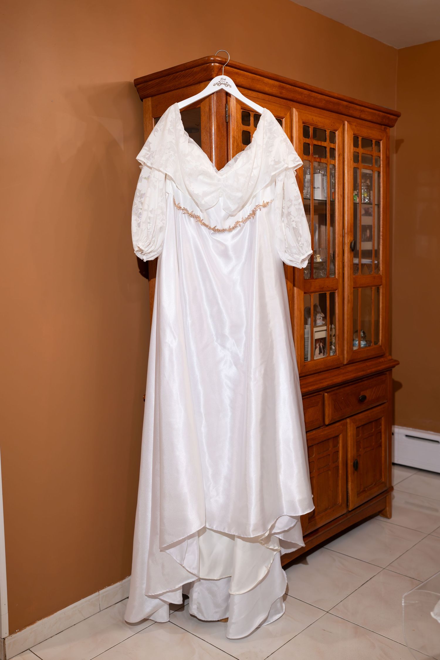 White satin off-shoulder wedding dress hanging on a wooden armoire in a warmly lit room.
