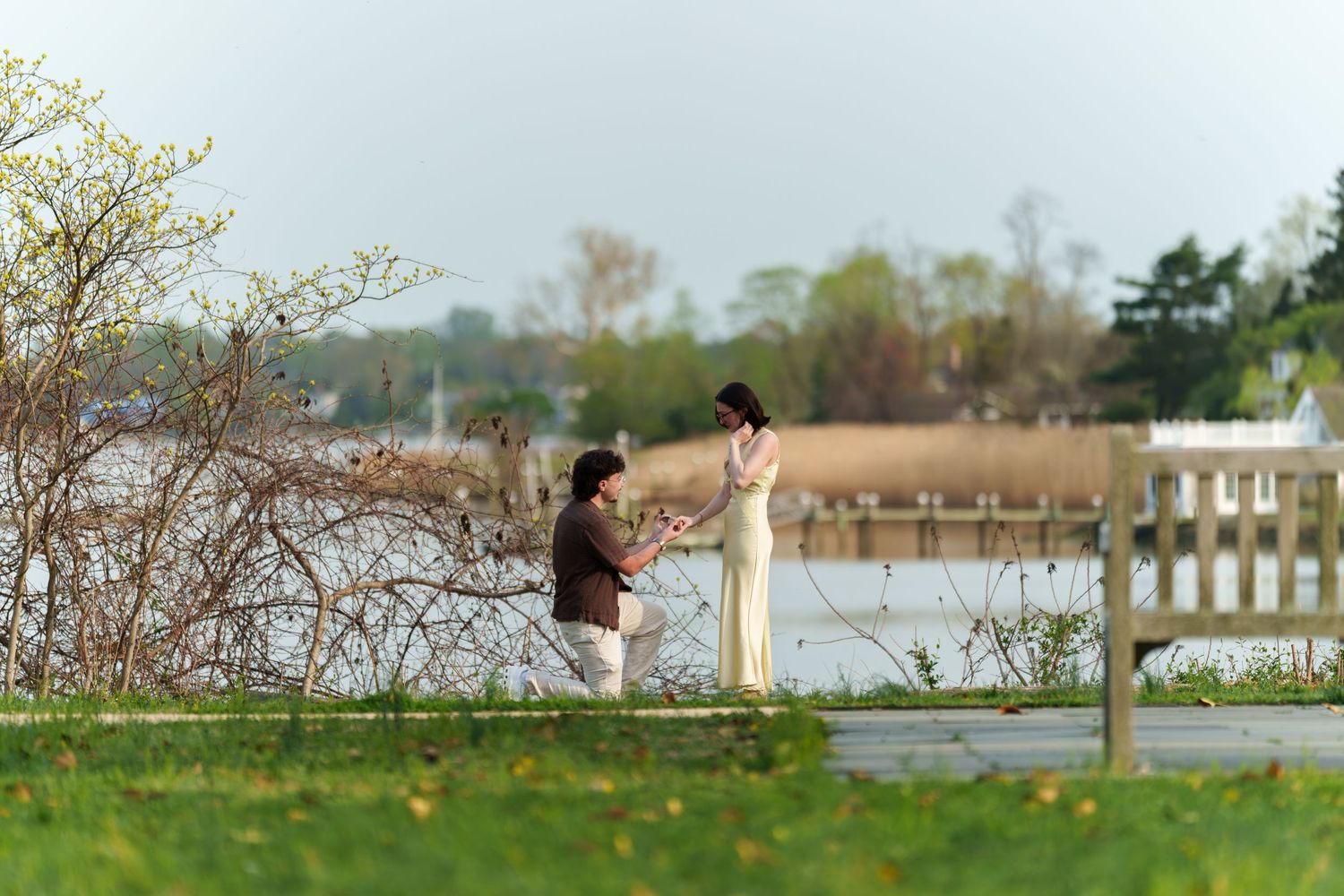 Man on one knee proposing to woman in elegant yellow dress near a peaceful lakeside path.
