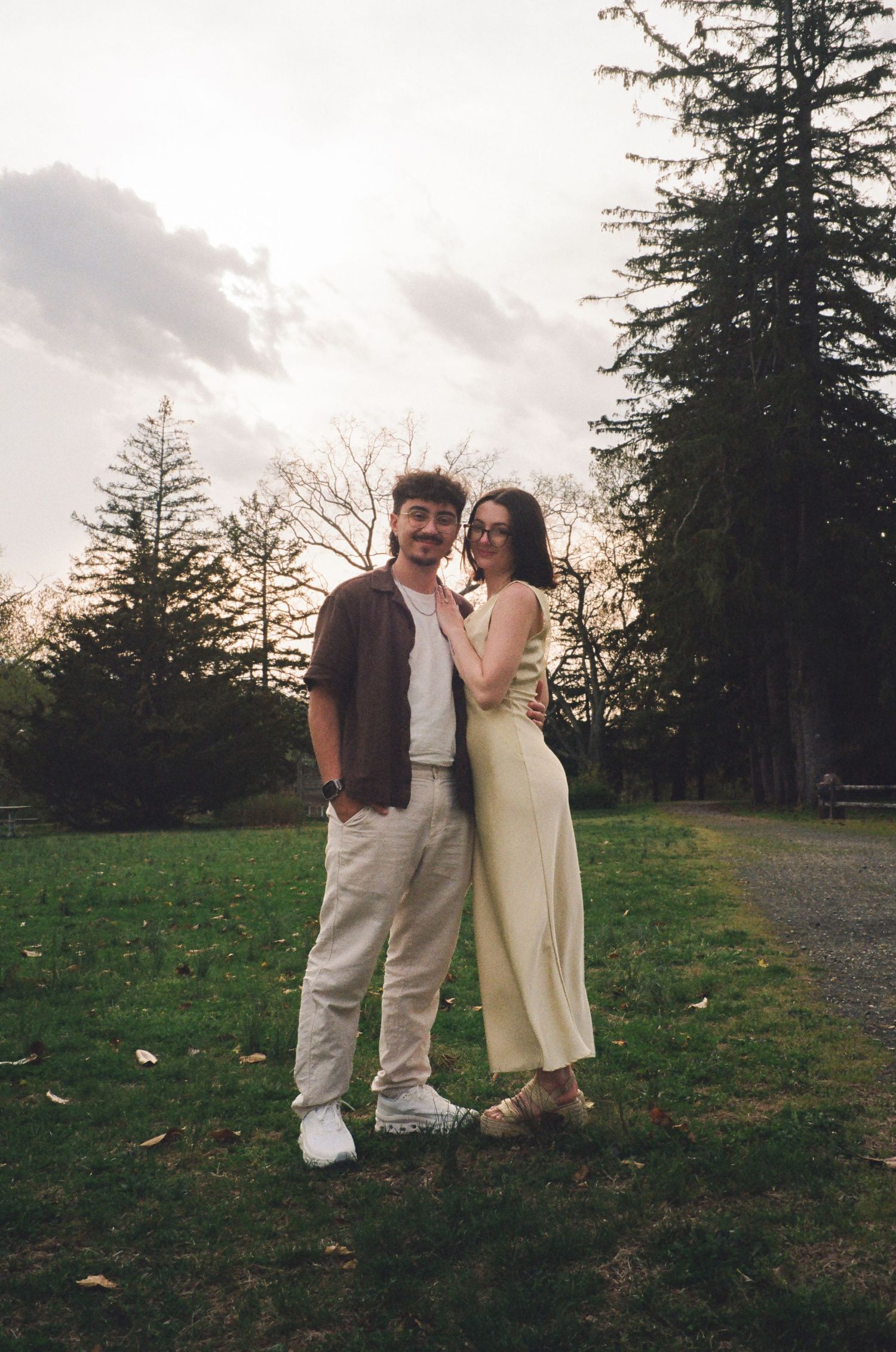 Happy couple posing together on a green park path surrounded by tall trees.