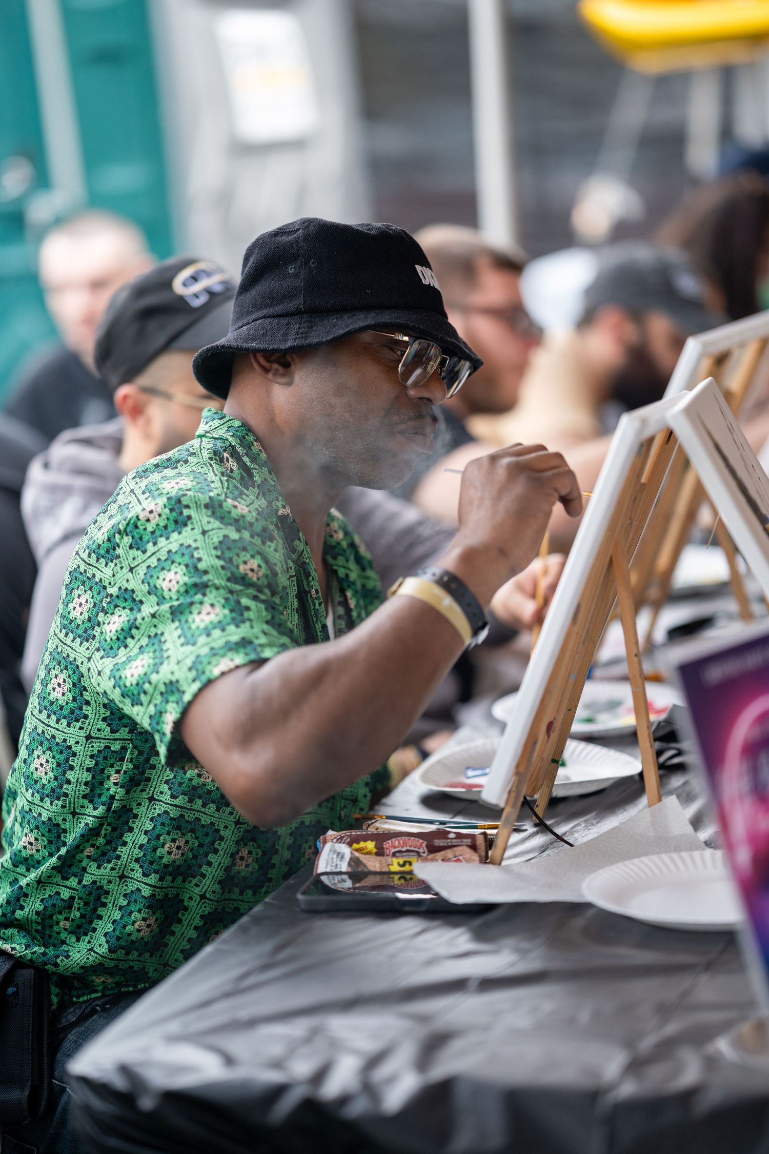 Man in green shirt and bucket hat painting at outdoor art event with other participants.