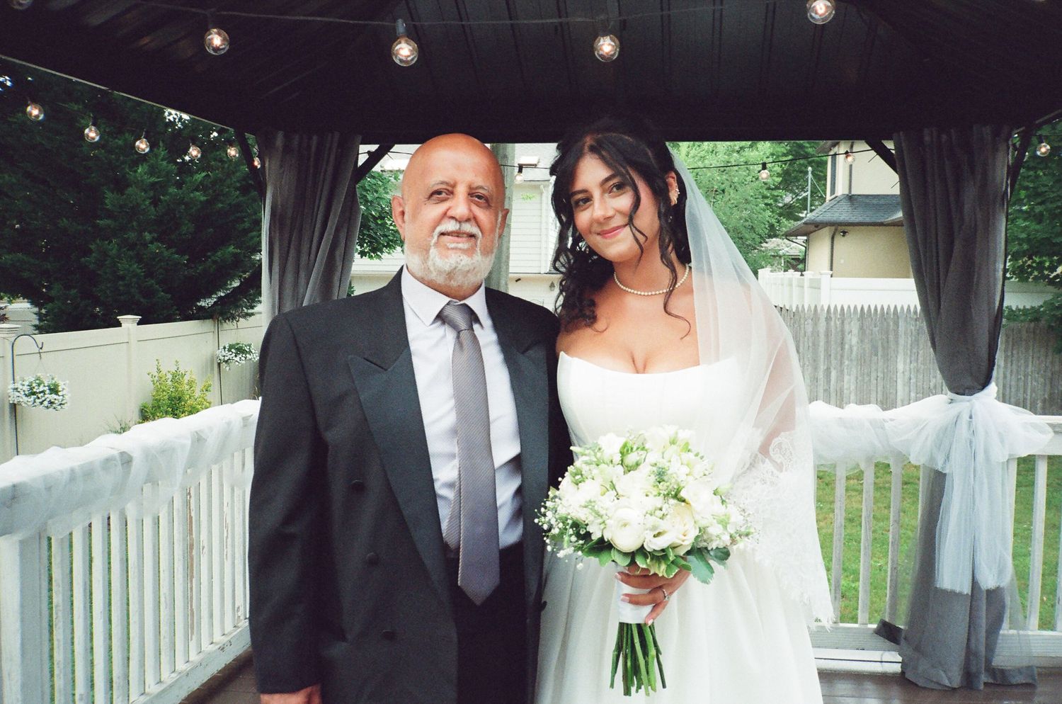 Bride in white gown holding white bouquet posing with elderly man at outdoor wedding.