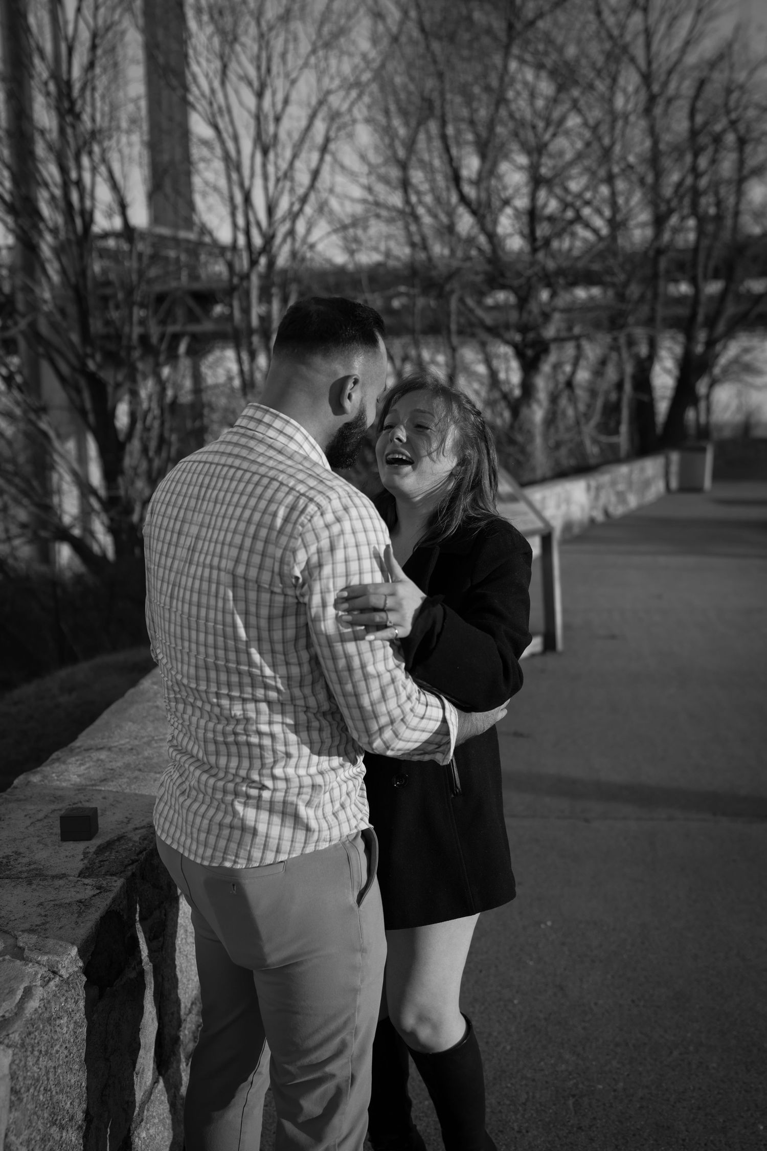 Black and white photo of happy couple embracing and laughing near stone wall after proposal.