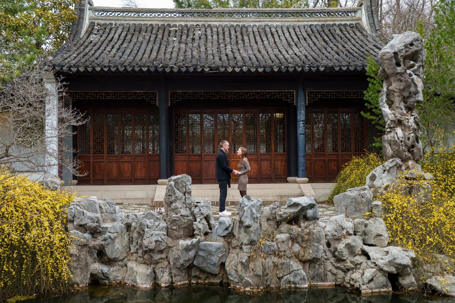 Couple standing on rocks beside a tranquil pond in front of a traditional Chinese pavilion with red doors.