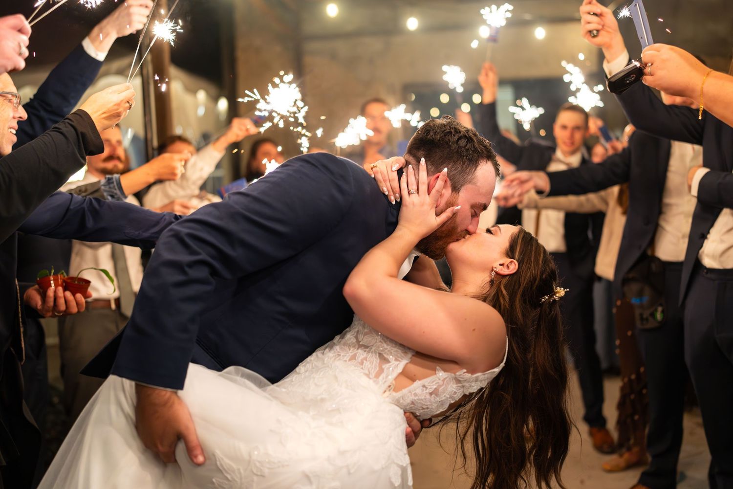 Groom dips bride for a passionate kiss surrounded by guests holding sparklers at wedding.