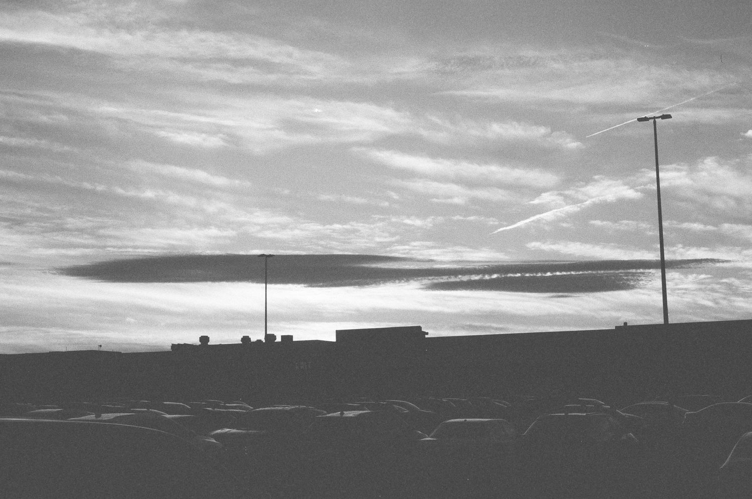 A dramatic cloud formation hovers over a parking lot and building silhouette at dusk in black and white.