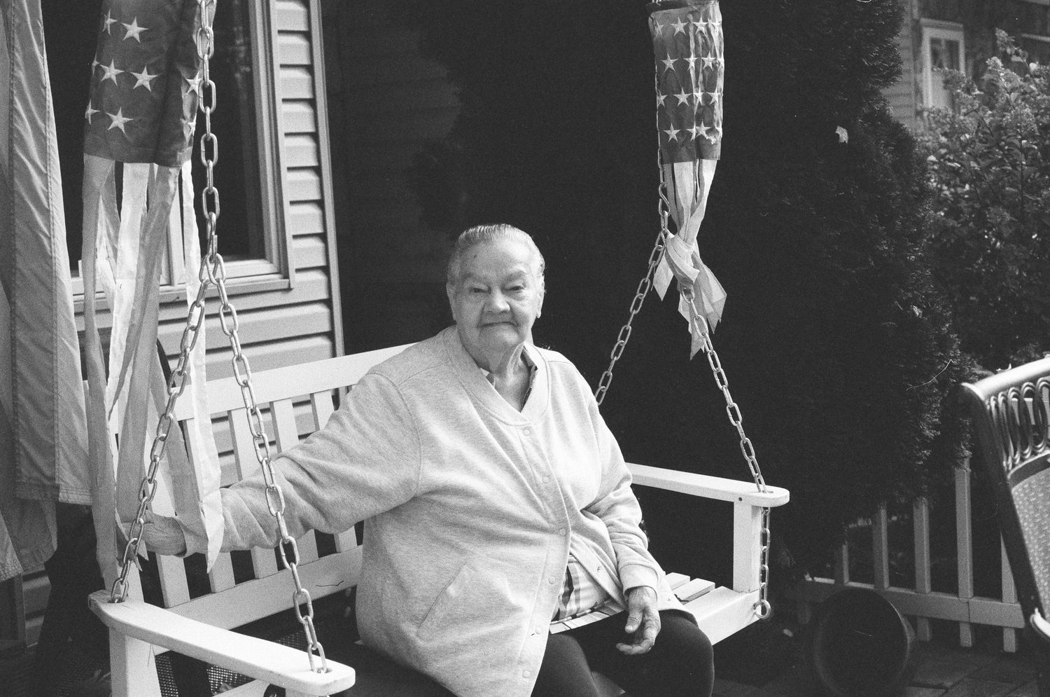 Elderly man sitting on a white porch swing with American flags in background.