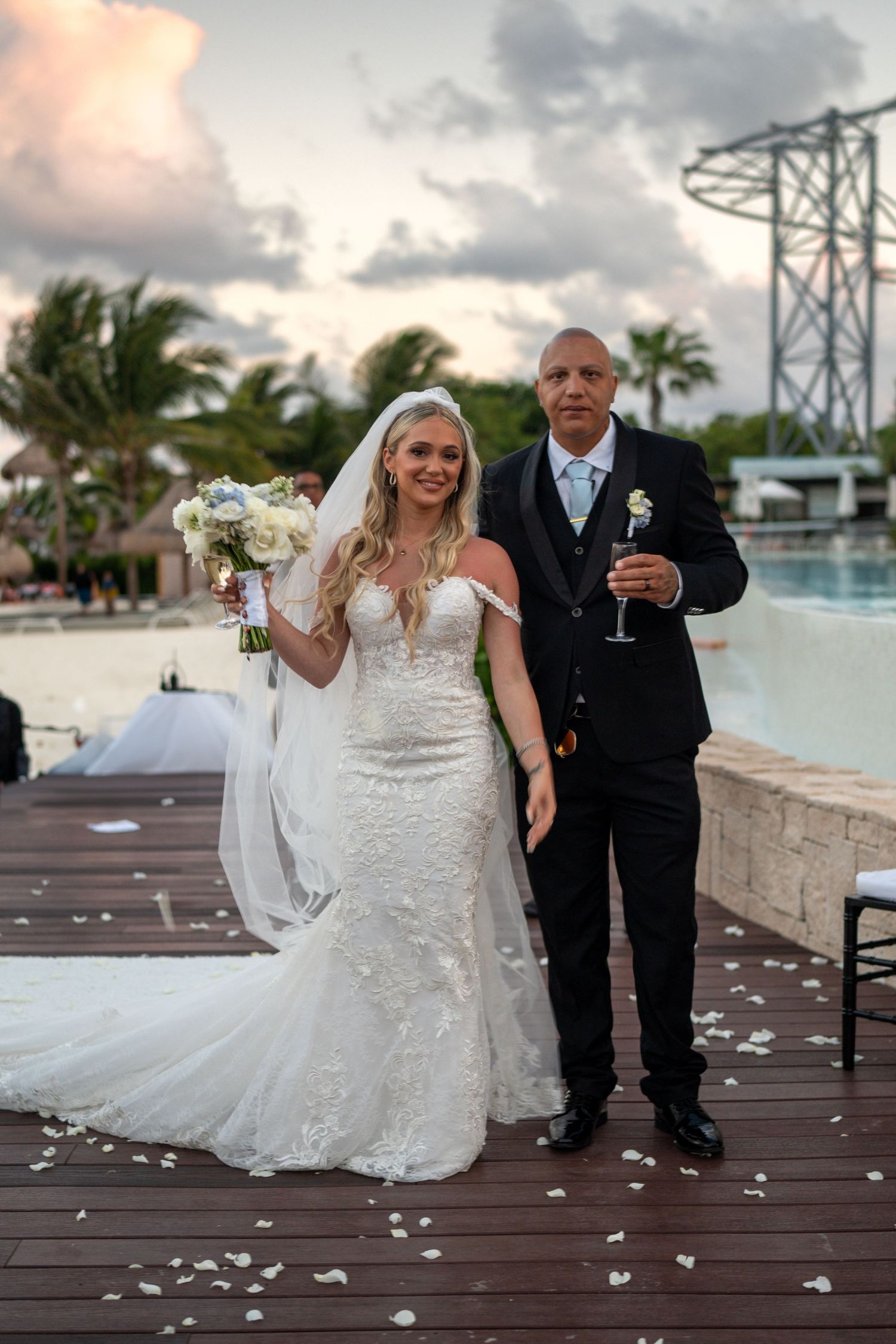 Smiling bride in lace gown holds white bouquet beside groom in black suit on rose petal-lined wedding aisle.