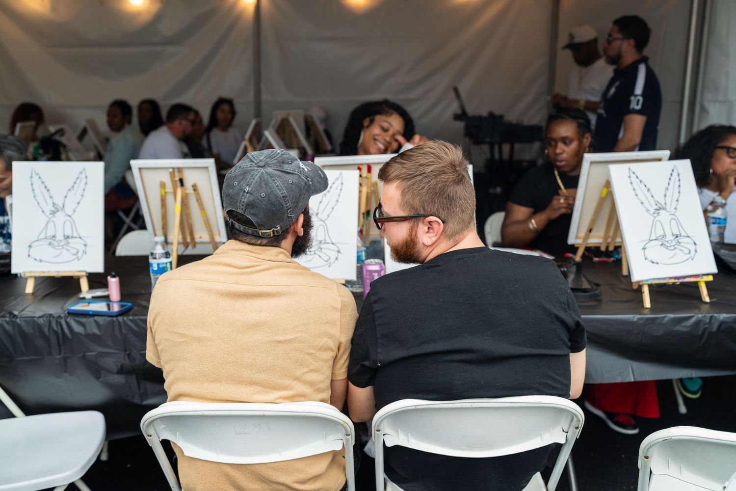 Two men sit together at a painting class with canvas easels inside a white outdoor tent.