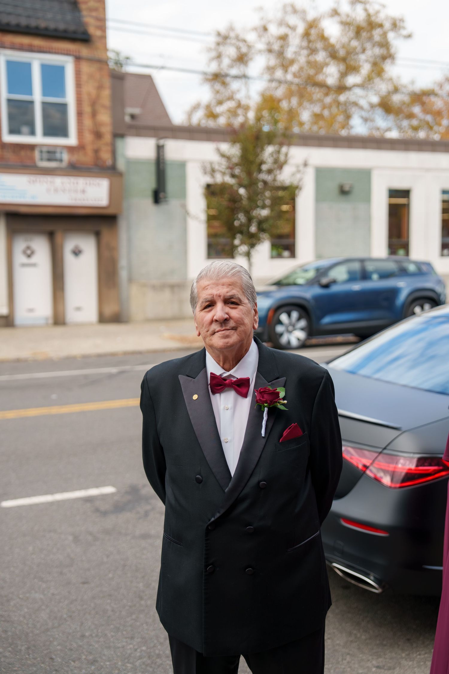 An older man in a black tuxedo with red bow tie and boutonniere stands on a city street.