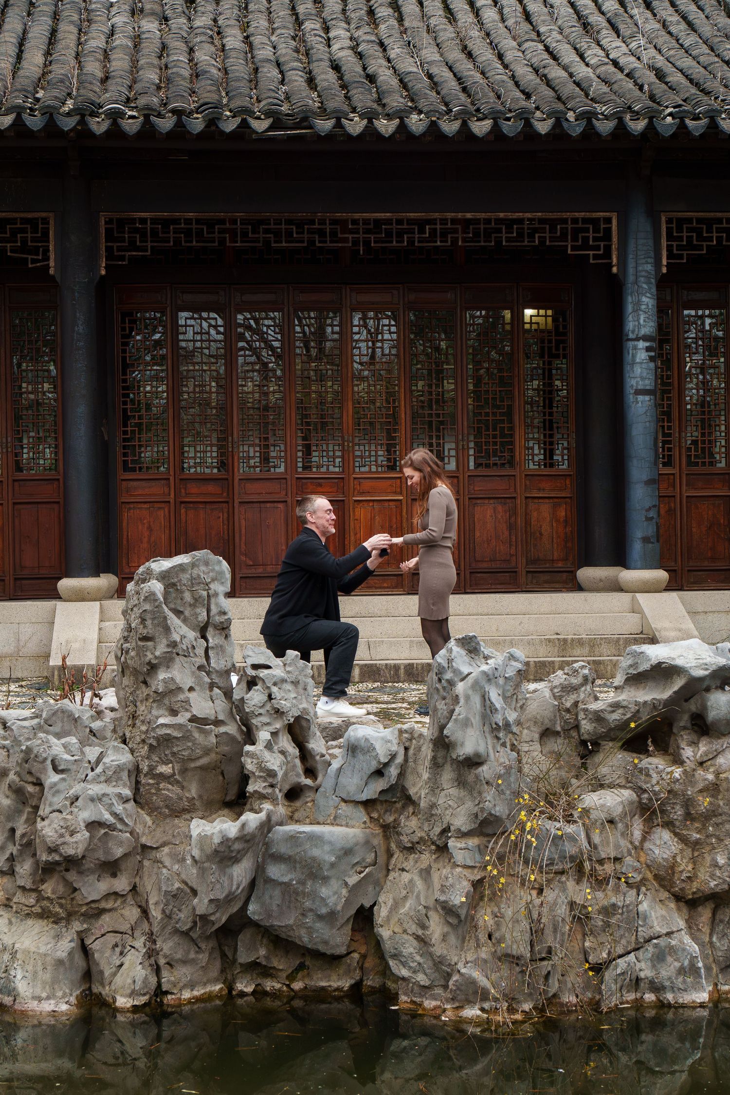 Man proposing on one knee to woman on rocks beside a pond at a Chinese garden.