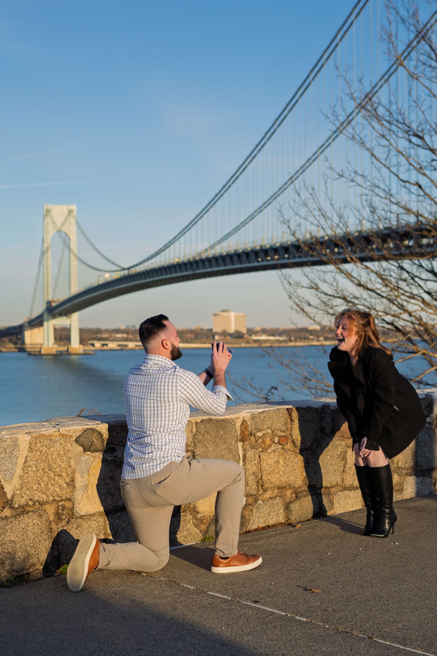Man proposing on one knee to woman near stone wall with Verrazzano-Narrows Bridge in background.