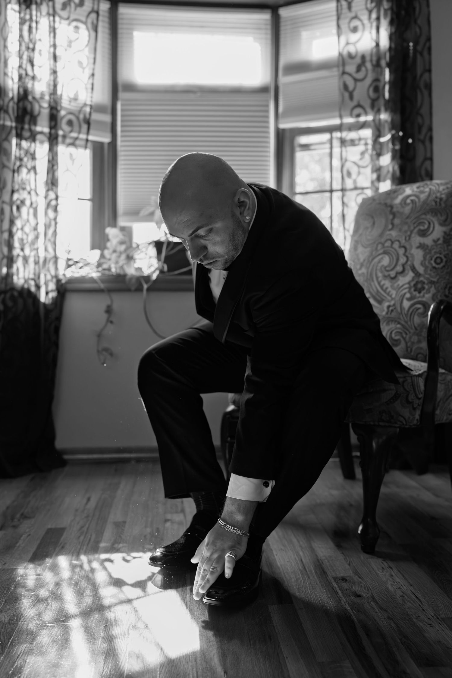 Black and white photo of a bald groom in a tuxedo polishing his shoes before his wedding.