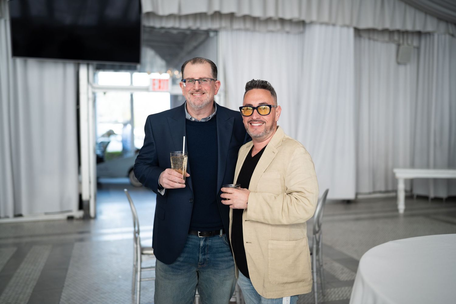 Two men smiling and holding drinks at an indoor event, one wearing yellow glasses.