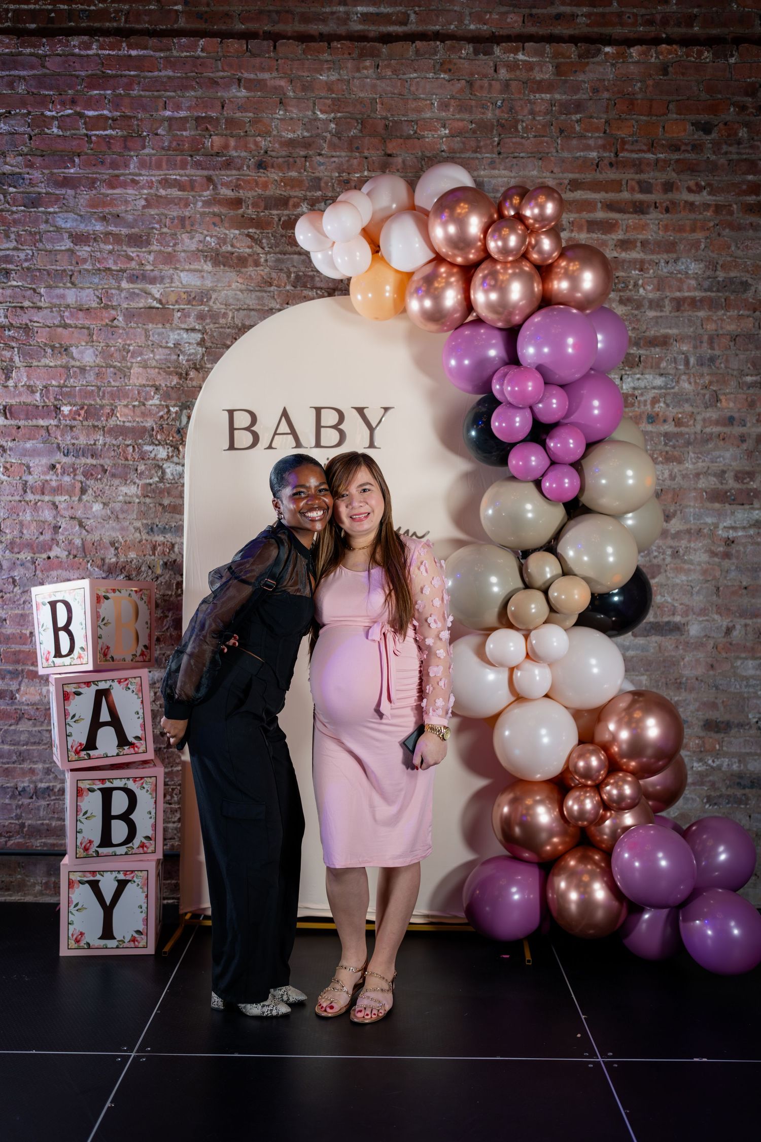 Two women smiling together at a baby shower, one visibly pregnant, in front of a colorful balloon arch backdrop.