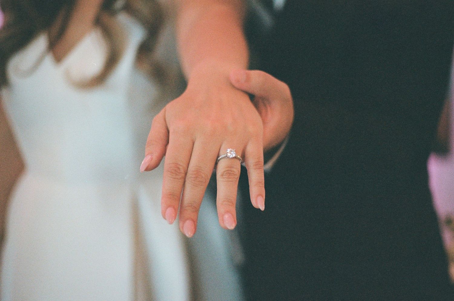 Close-up of a couple holding hands displaying a diamond engagement ring, wearing wedding attire.