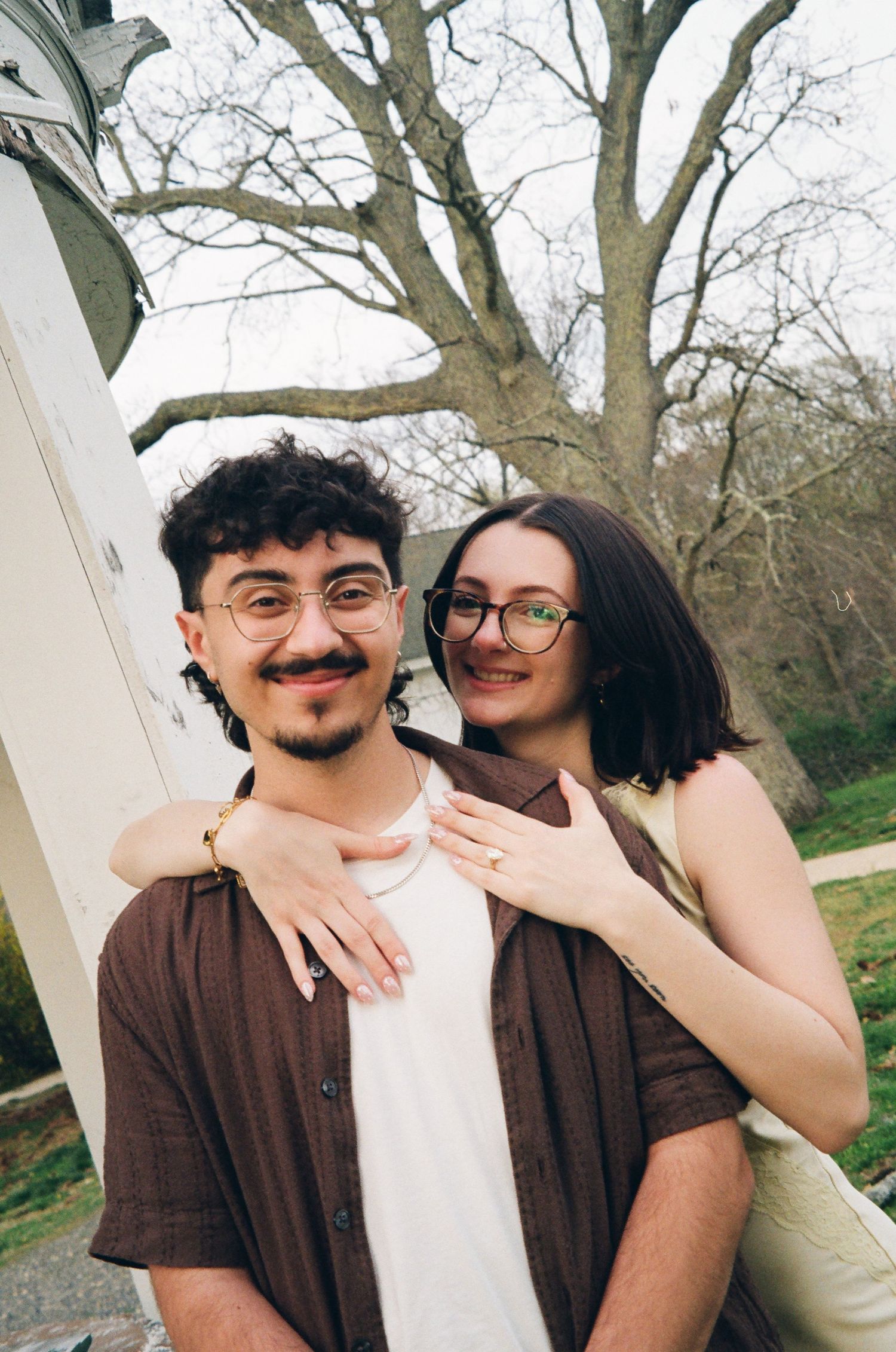 Couple posing near white columns, woman in yellow dress hugging man in brown shirt from behind.