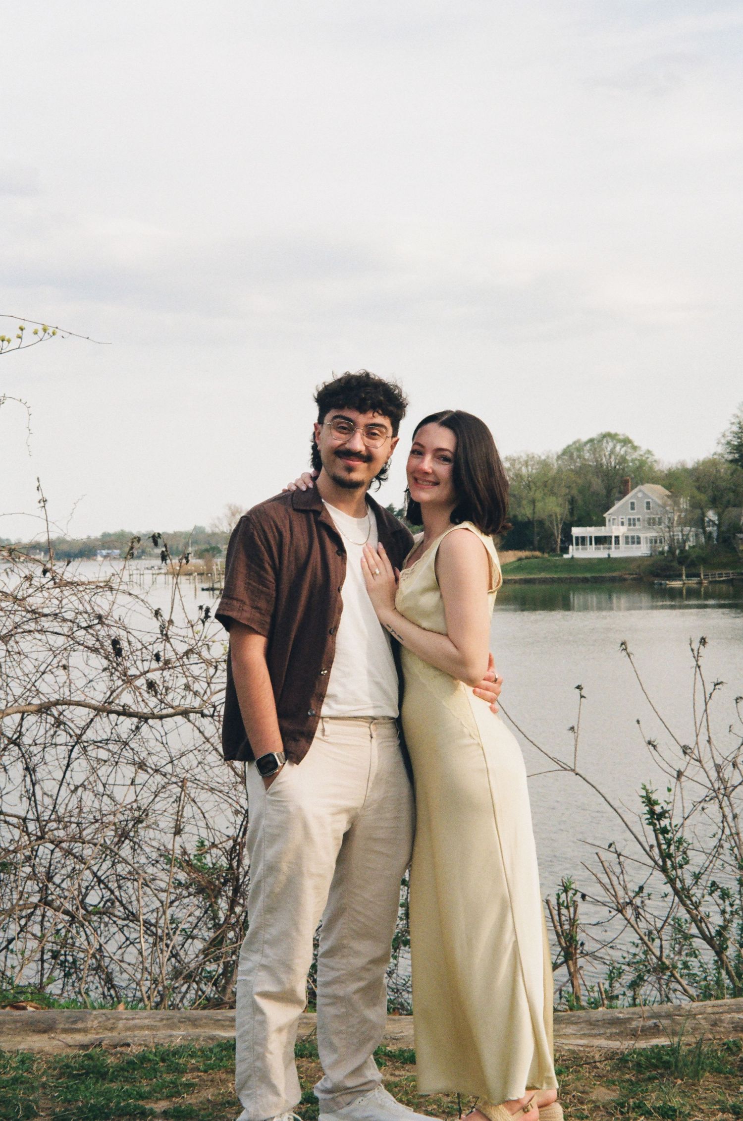 Couple posing together by a lakeside with a white house and trees reflected in the calm water.