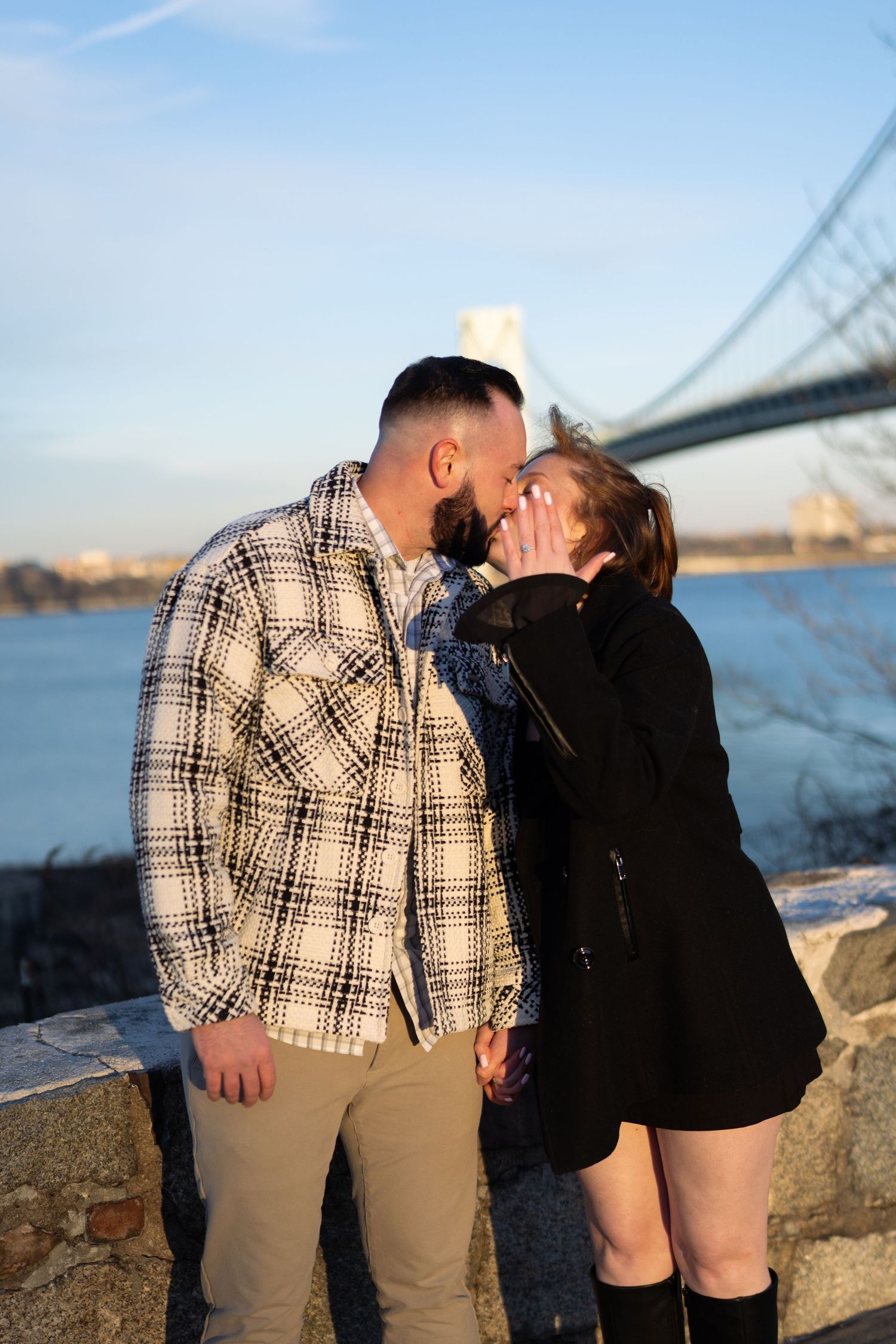 Couple sharing a kiss outdoors near the Verrazzano-Narrows Bridge during golden hour sunset.