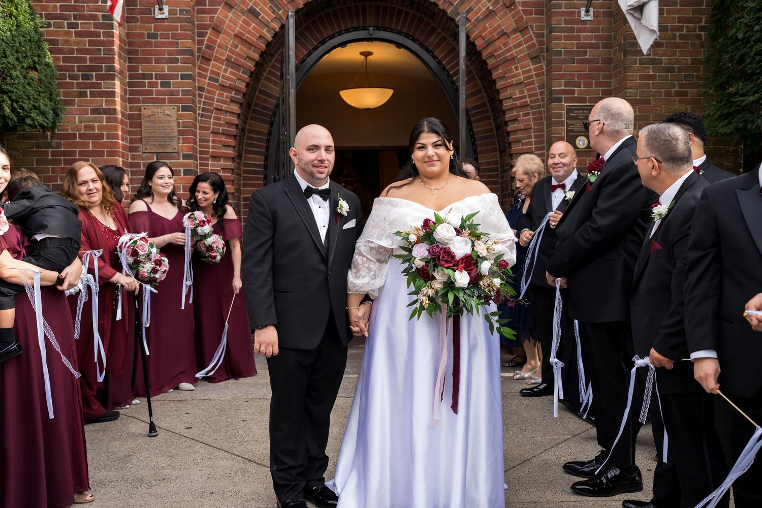 Bride and groom exit brick church greeted by bridesmaids in burgundy dresses and groomsmen.