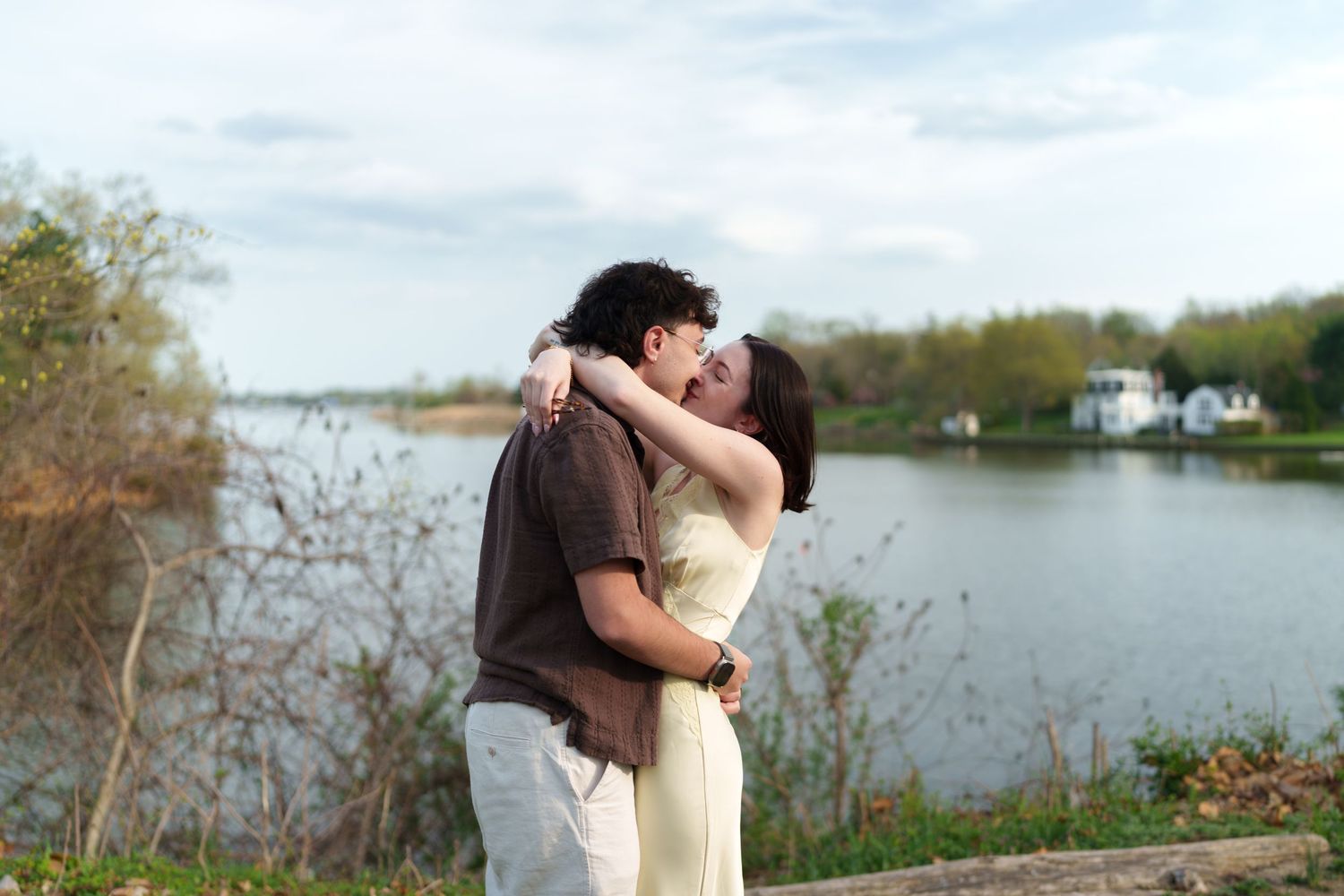 Romantic couple sharing a kiss outdoors near a peaceful lake on a cloudy day.