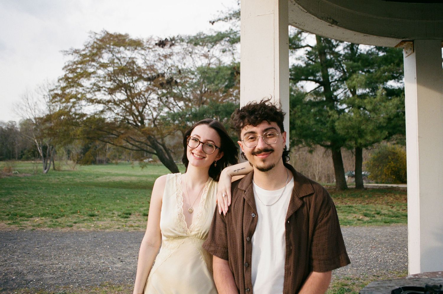 Couple smiling under a white column, woman in yellow dress with glasses, man in brown shirt.